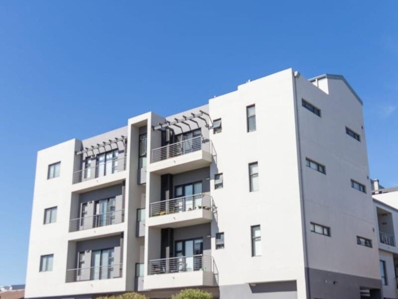 A modern multi-story apartment building featuring private balconies with metal railings, pergola structures, and a paved street-level entrance.