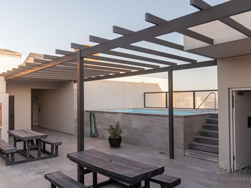 A modern rooftop terrace featuring a built-in swimming pool, pergola, glass balustrade, and a built-in braai area with tiled flooring.