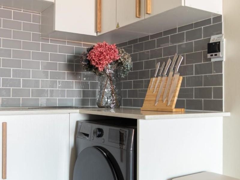 Modern kitchen area featuring white cabinetry with timber handles, grey subway tile backsplash, and a built-in washing machine under a stone countertop.