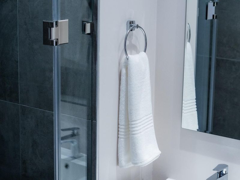 A modern bathroom detail featuring dark wall tiling, a glass shower screen, a wall-mounted towel ring, and a sleek basin mixer tap.