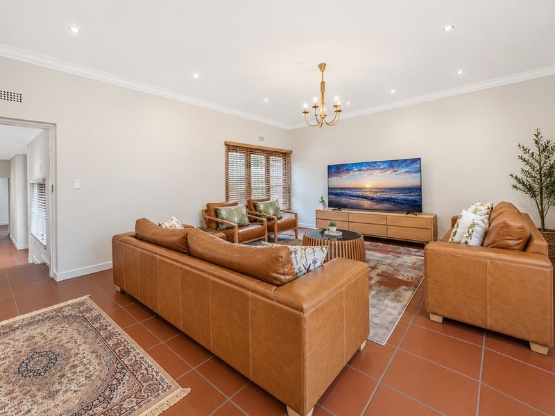 A well-maintained living room featuring tiled flooring, recessed downlights, a central chandelier, and timber venetian blinds.