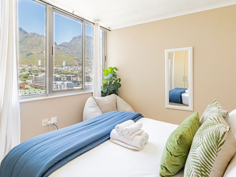 A bright bedroom featuring large windows with expansive mountain and city views, neutral wall finishes, and clean, well-maintained architectural elements.