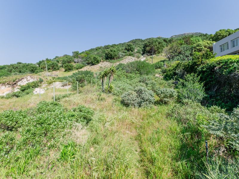 A view of a sloped, undeveloped residential plot featuring natural vegetation, shrubs, and perimeter wire fencing.