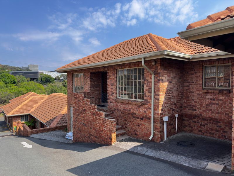 Exterior view of a face-brick townhouse featuring a terracotta tiled roof, grid-pattern windows, and a paved driveway entrance.