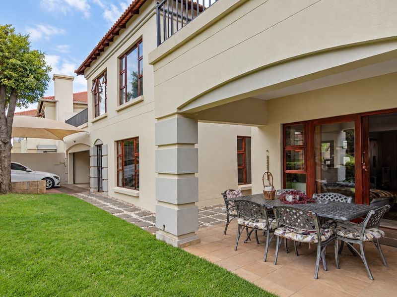 A well-maintained rear exterior view featuring a covered tiled patio, timber-framed windows, and a balcony overlooking a landscaped garden area.