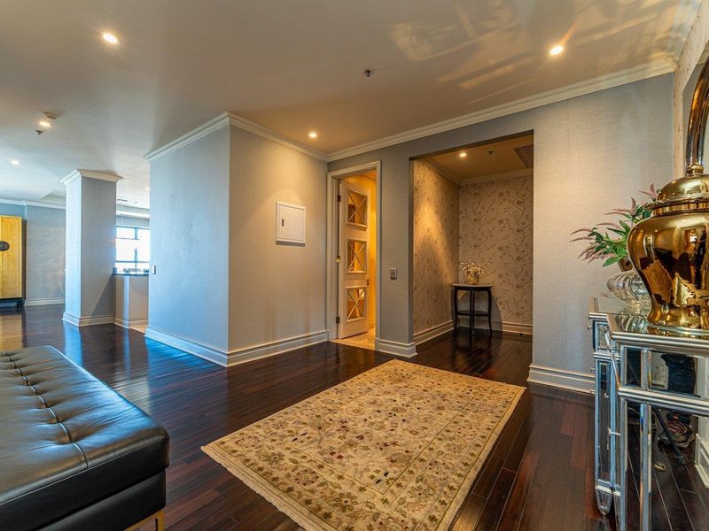 A spacious and well-lit hallway featuring dark hardwood flooring, elegant crown molding, and decorative textured wallpaper, leading to multiple interior rooms.