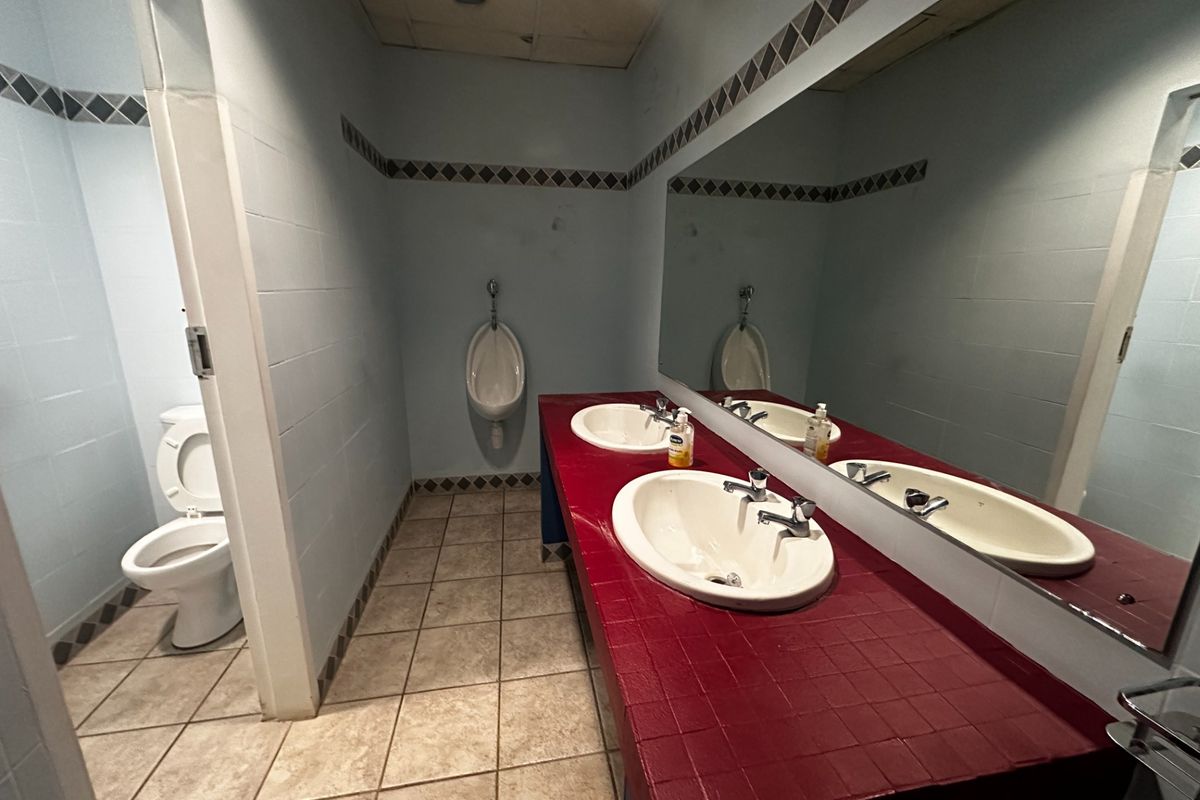 Commercial restroom featuring a dual vanity with tiled countertop, wall-mounted urinal, and a separate toilet stall with tiled flooring.