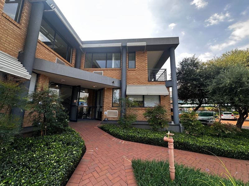 A well-maintained commercial building exterior featuring a brick facade, paved entrance walkway, and a balcony, surrounded by landscaped greenery.