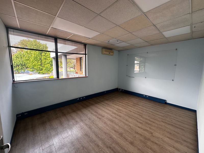 A vacant office space featuring laminate flooring, a suspended ceiling, wall-mounted air conditioning, and a glass whiteboard, offering a functional workspace environment.