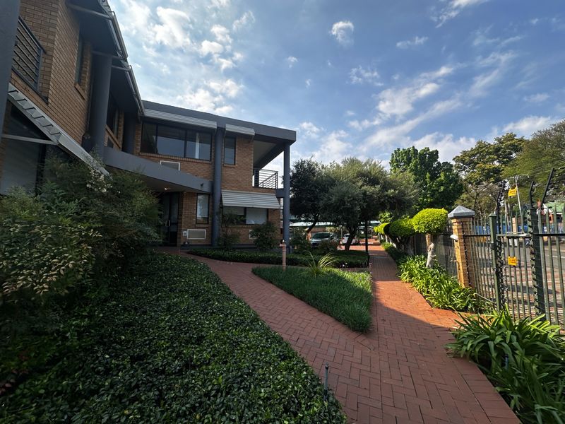 Front exterior view of a commercial property featuring a brick facade, paved walkway, landscaped garden, and secure perimeter fencing.