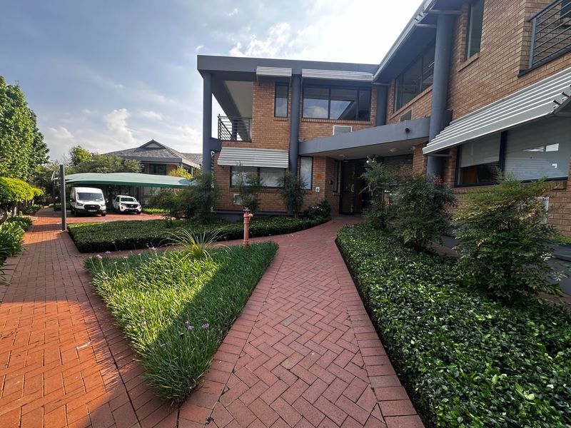 Front exterior view of a commercial brick building featuring a paved walkway, landscaped garden beds, and a covered parking area.