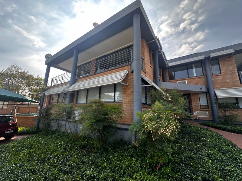 A multi-story commercial building featuring a brick facade, covered balcony with metal railings, and a paved entrance walkway surrounded by landscaped greenery.