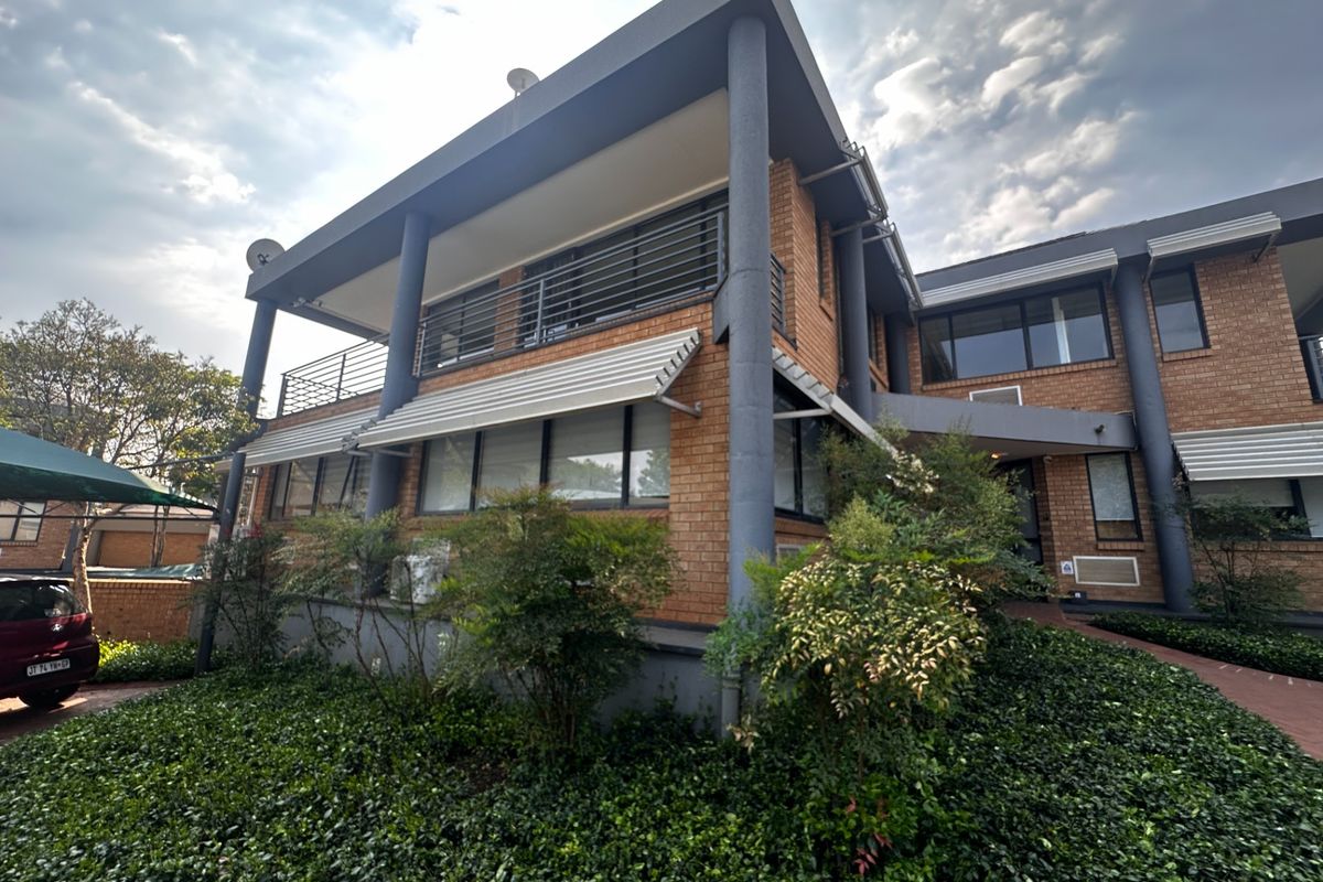A multi-story commercial building featuring a brick facade, covered balcony with metal railings, and a paved entrance walkway surrounded by landscaped greenery.