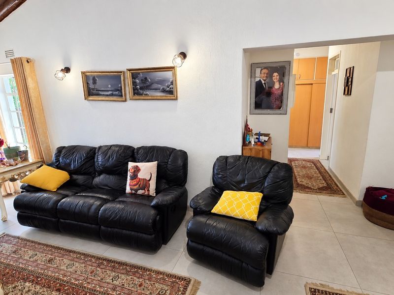 A living room featuring tiled floors, exposed timber ceiling beams, and wall-mounted lighting, with an adjacent hallway leading to built-in storage cupboards.
