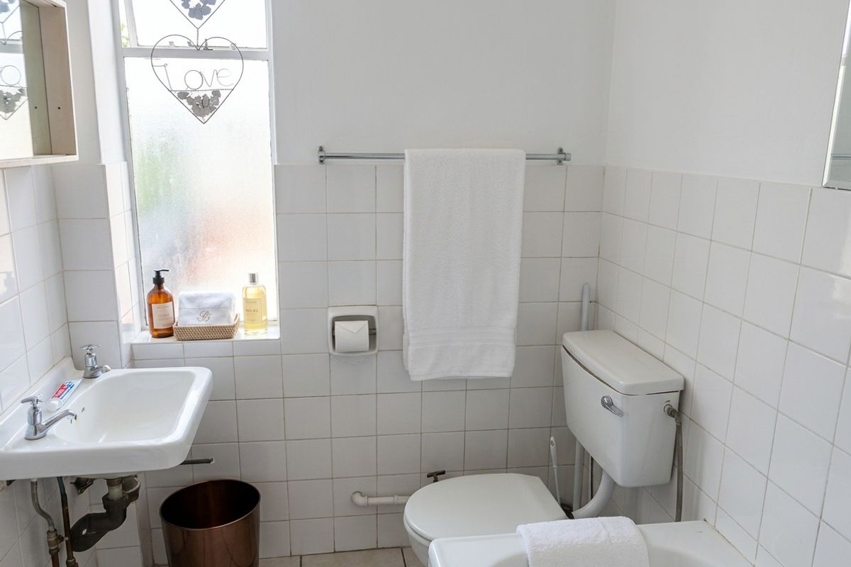 A functional bathroom featuring white wall tiles, a wall-mounted basin, and a standard toilet with a frosted window for natural light.