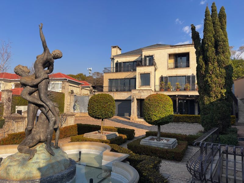 A grand multi-story residence featuring a paved driveway, ornate fountain, manicured topiary, and wrought iron balcony railings.
