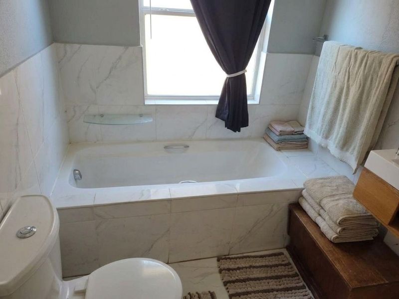 A functional bathroom featuring a built-in bathtub with marble-look tile surround, a toilet, and a window providing natural light.