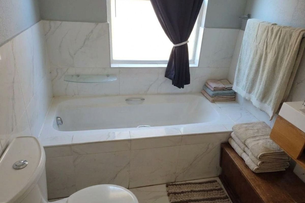 A functional bathroom featuring a built-in bathtub with marble-look tile surround, a toilet, and a window providing natural light.