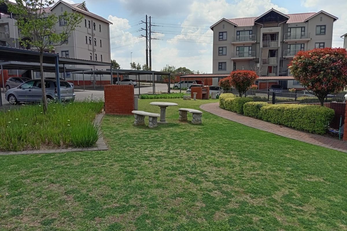A well-maintained communal garden area featuring a brick braai, paved pathways, and established landscaping within an apartment complex setting.
