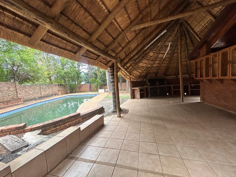 Spacious covered patio with a thatched roof, built-in braai, and wooden cabinetry, overlooking a private swimming pool and garden area.