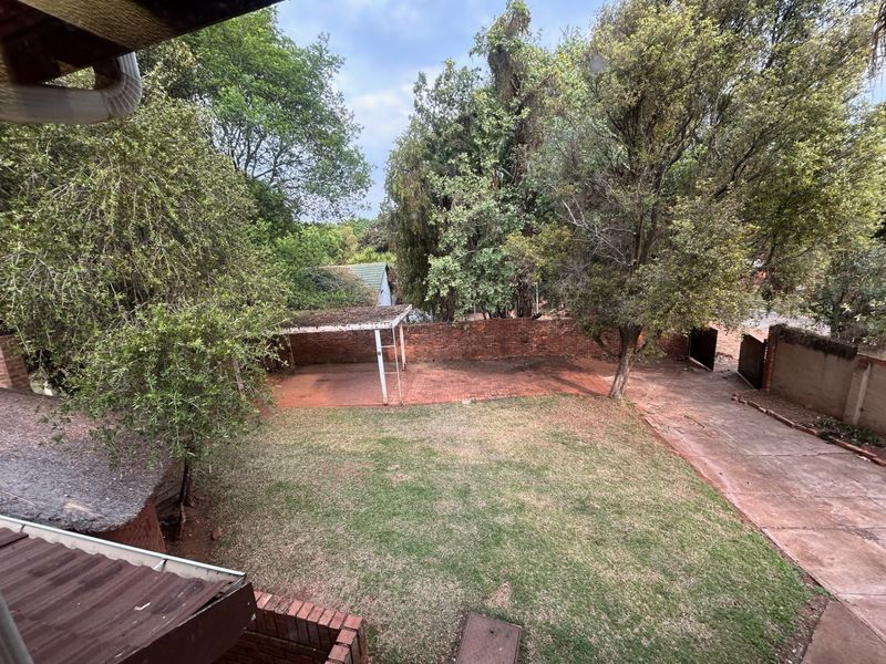 A rear garden view featuring a lawn, mature trees, a brick boundary wall, and a paved driveway with a covered carport structure.