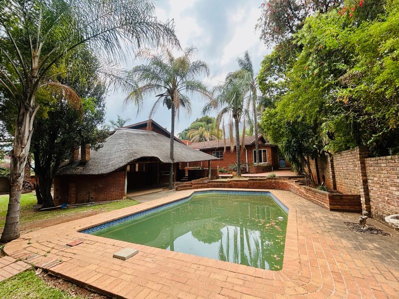 Outdoor pool area featuring a swimming pool, brick paving, and a thatched lapa structure set within a mature garden.