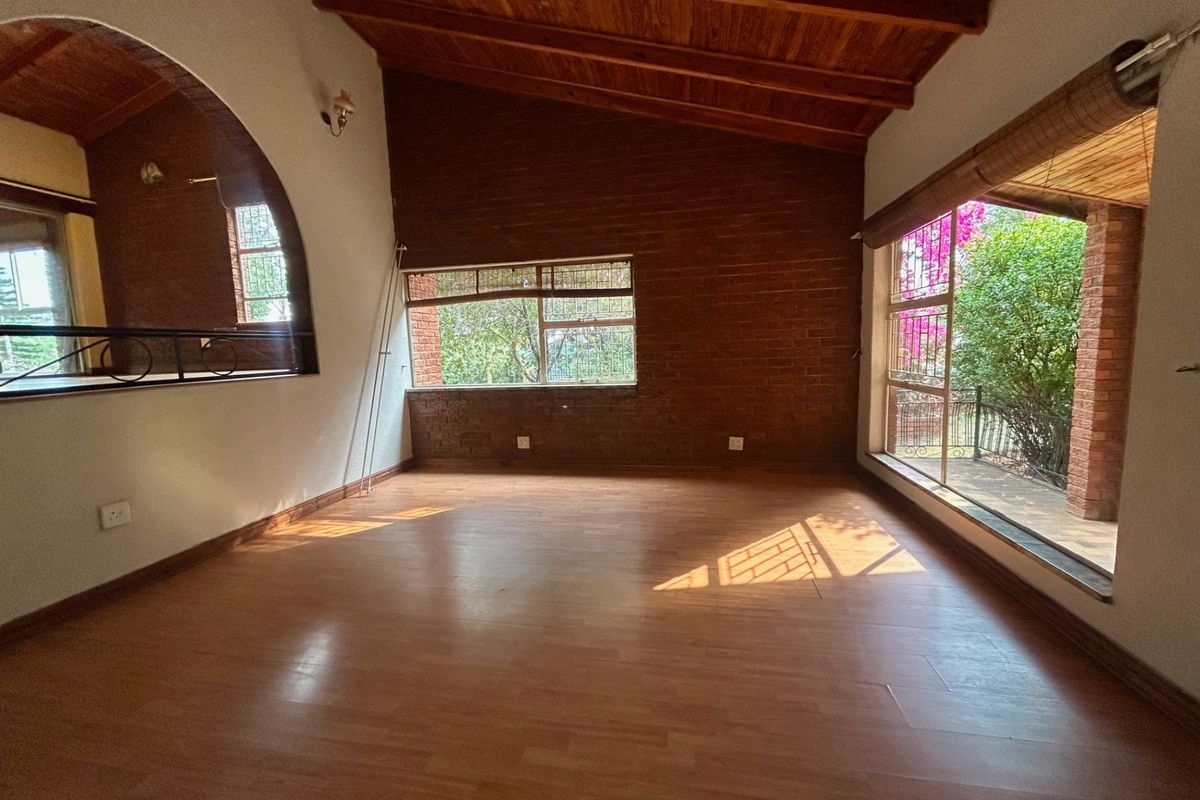Spacious living area featuring exposed timber beam ceilings, a rustic brick feature wall, and large windows providing natural light and garden views.
