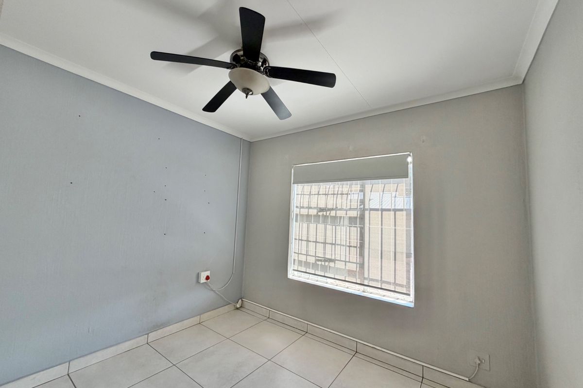 An empty bedroom featuring tiled flooring, a ceiling fan, and a window fitted with security bars and a roller blind.