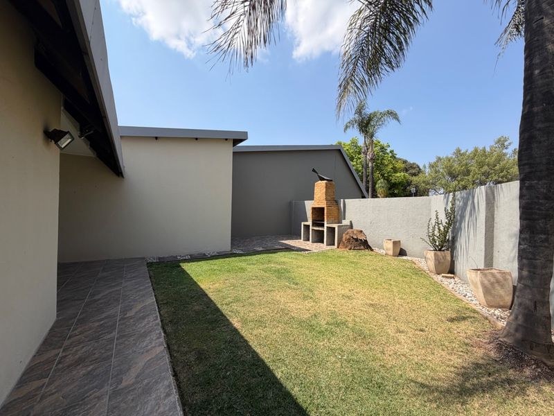 A private rear garden featuring a tiled patio area, a built-in brick braai, and a well-maintained lawn enclosed by boundary walls.