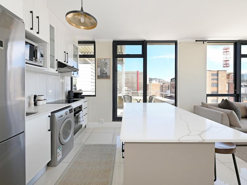 Modern open-plan kitchen and living area featuring sleek white cabinetry, stone island countertop, integrated appliances, and large sliding doors leading to a balcony.
