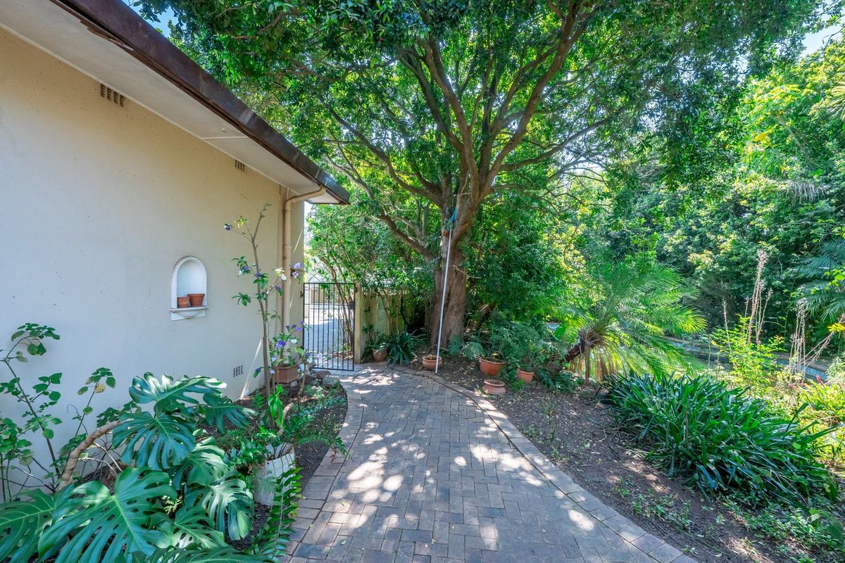 A peaceful rear garden path featuring paved walkways, mature trees, and a charming recessed wall niche, leading to a secure wrought iron gate.