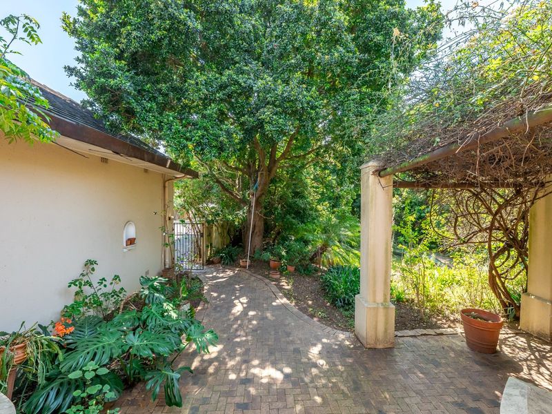 A tranquil garden area featuring brick paving, a shaded pergola structure with concrete pillars, and mature trees providing natural shade.