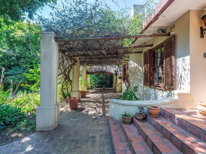 A covered patio area featuring a rustic pergola, brick paving, and terracotta steps, leading into a lush, mature garden setting.
