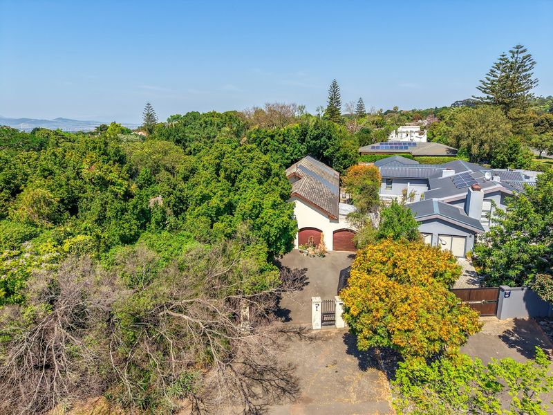 Aerial view of the property showcasing the main residence, double garage, paved driveway, gated entrance, and surrounding mature garden landscape.