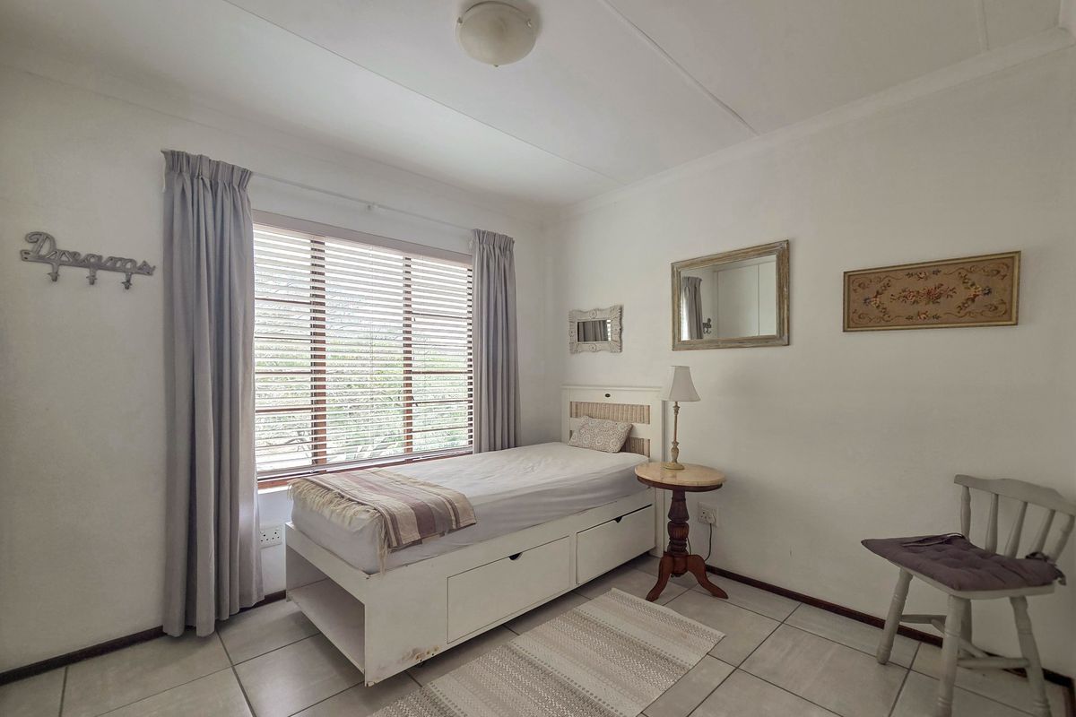 A functional bedroom featuring tiled flooring, neutral walls, and a large window fitted with venetian blinds, providing natural light to the space.