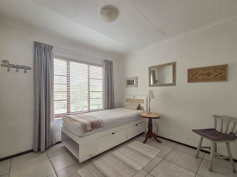 A functional bedroom featuring tiled flooring, neutral walls, and a large window fitted with venetian blinds, providing natural light to the space.