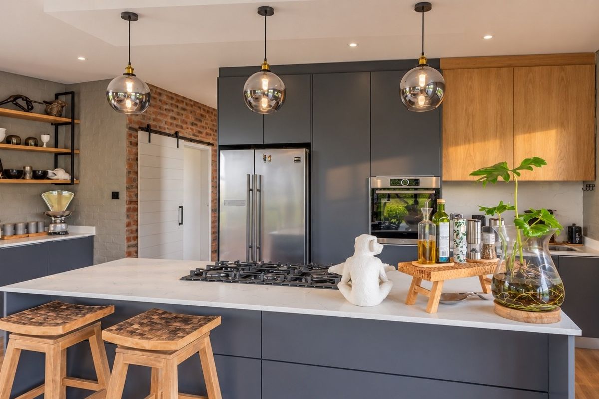 Modern kitchen featuring dark cabinetry, stone countertops, a central island with a gas hob, and stylish pendant lighting against an exposed brick feature wall.