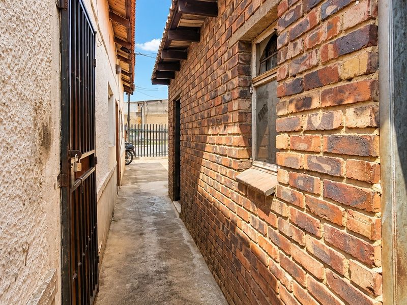 A narrow exterior side passage featuring exposed brick walls, a concrete walkway, and a metal security gate, showing signs of age and weathering.