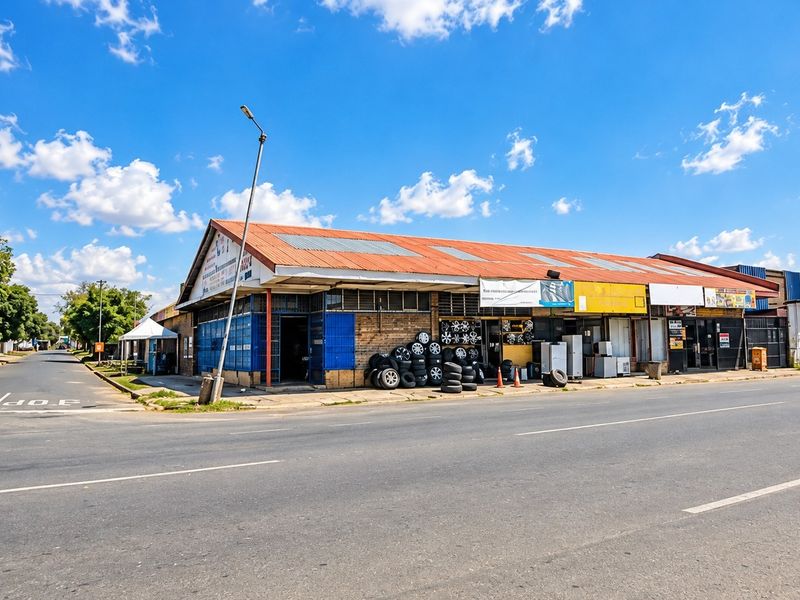 Street-level view of a commercial property featuring a corrugated metal roof, brick facade, and multiple storefront access points along a main road.