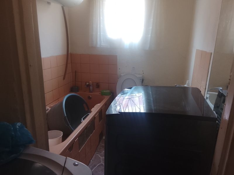 A dated bathroom featuring a built-in bathtub with tiled surround, toilet, and window, requiring renovation to update the finishes and fixtures.