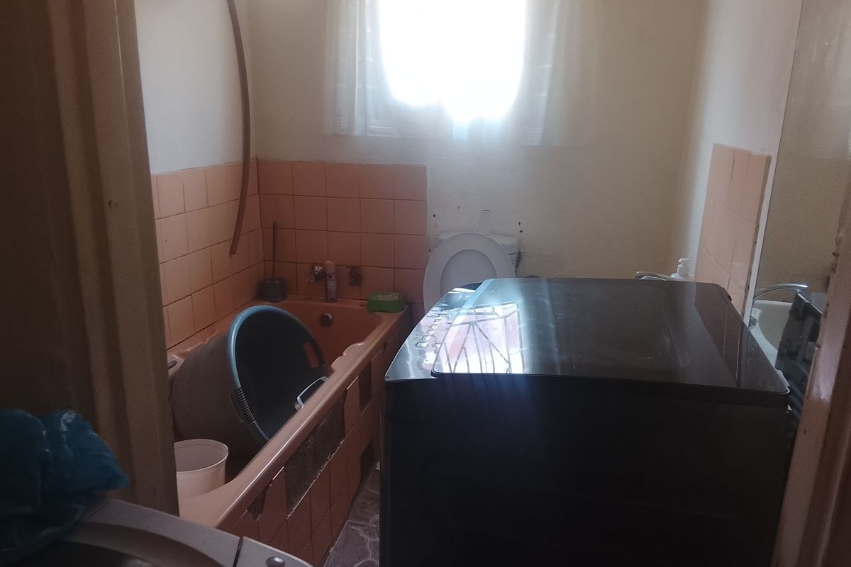 A dated bathroom featuring a built-in bathtub with tiled surround, toilet, and window, requiring renovation to update the finishes and fixtures.