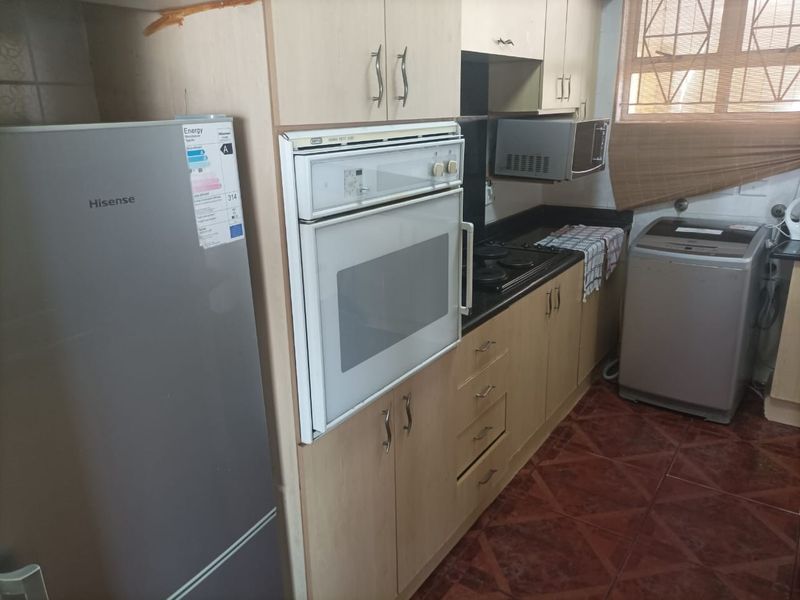 A compact kitchen featuring light wood-finish cabinetry, built-in oven and hob, dark countertops, and tiled flooring, with visible wear on the ceiling trim.