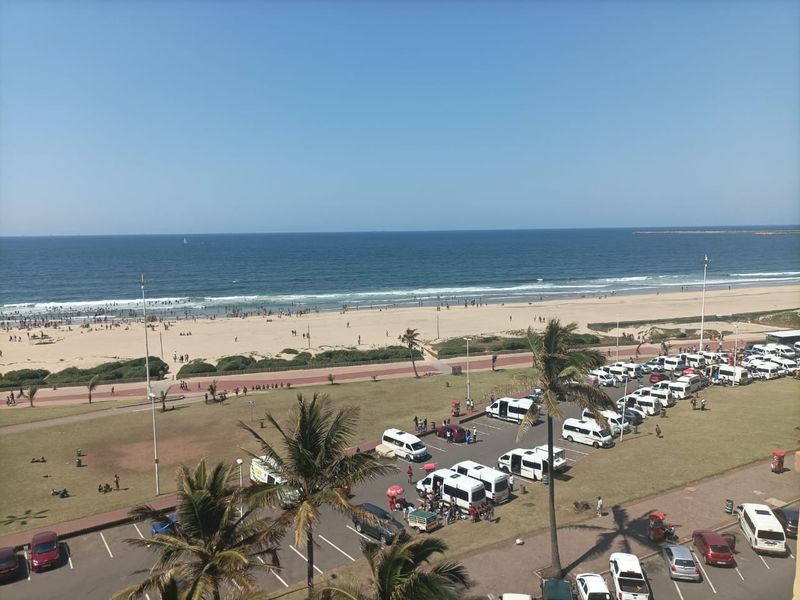 Elevated coastal view overlooking the beach, promenade, and public parking area.
