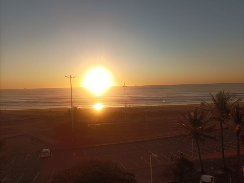 A scenic elevated view of the coastline at sunset, overlooking a parking area and the ocean horizon.