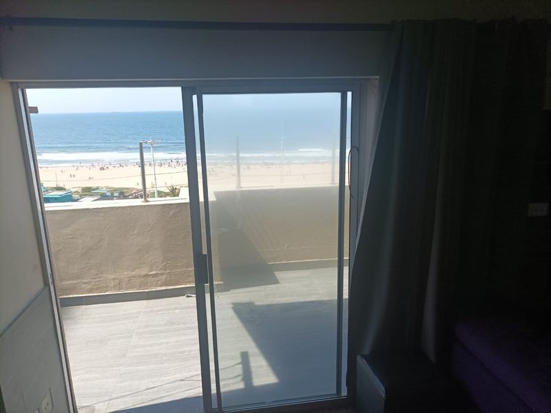 A view of the beach and ocean from an interior room through a sliding glass door leading to a tiled balcony.
