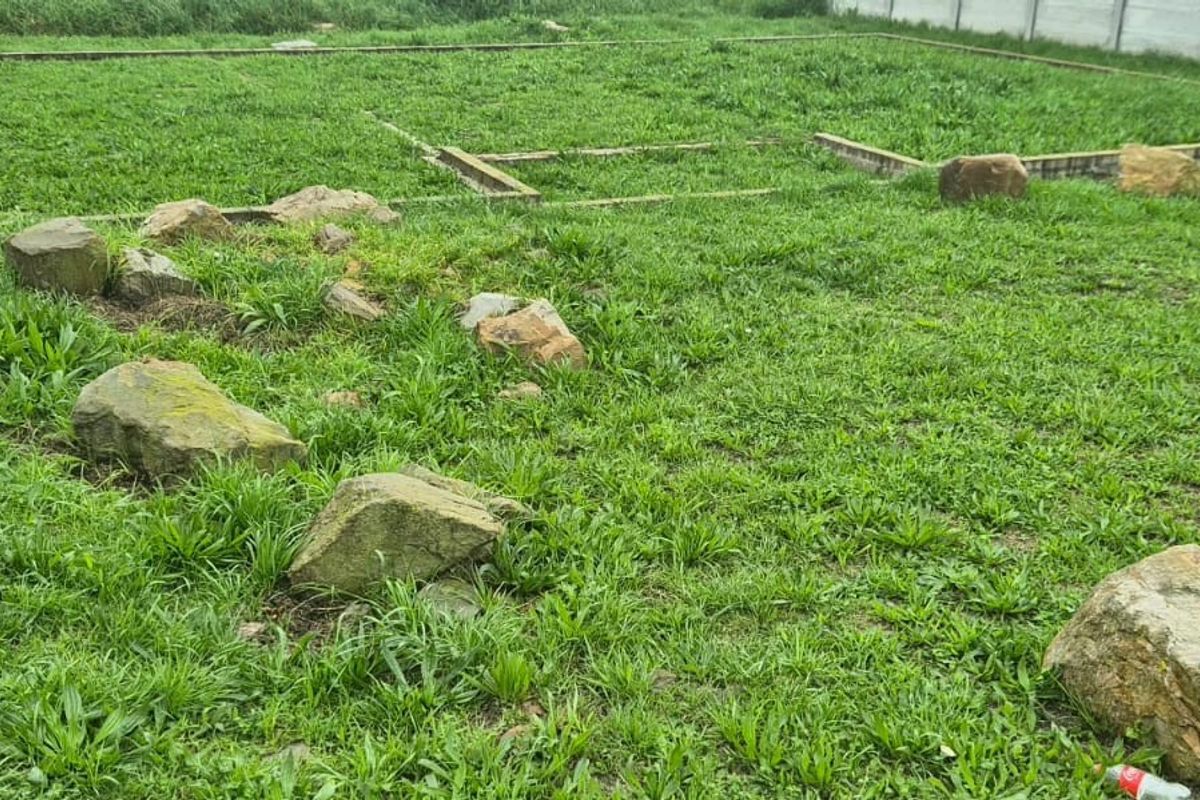 A vacant residential plot featuring established concrete boundary walls and visible foundation footings, surrounded by overgrown grass and natural rock features.