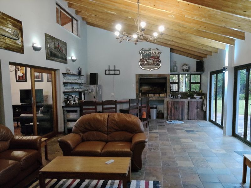 Spacious open-plan living area featuring exposed timber rafters, stone tile flooring, a built-in bar, and an indoor braai, with direct access to the garden.