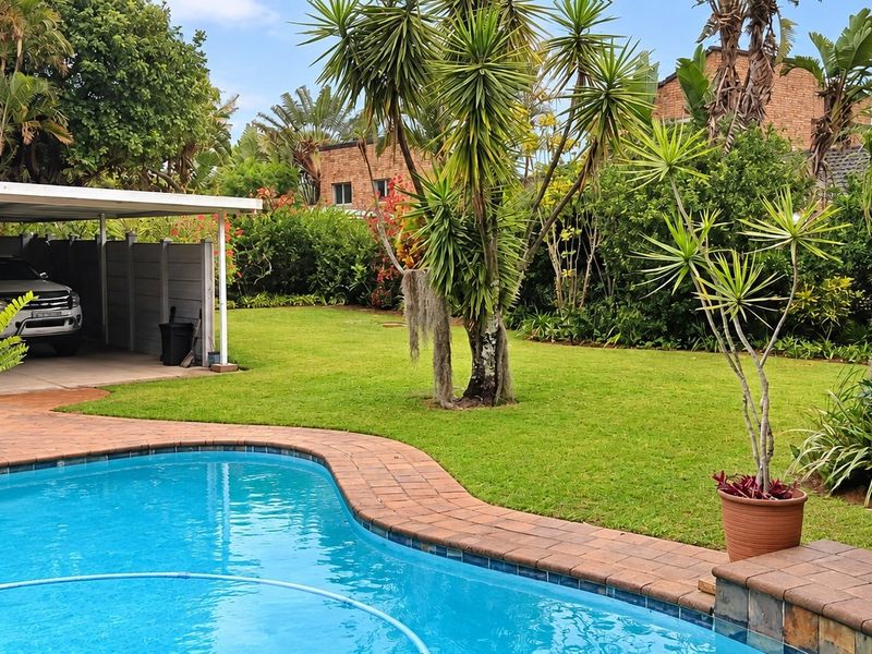 A well-maintained outdoor area featuring a swimming pool with brick coping, a lush garden, and an attached covered carport.