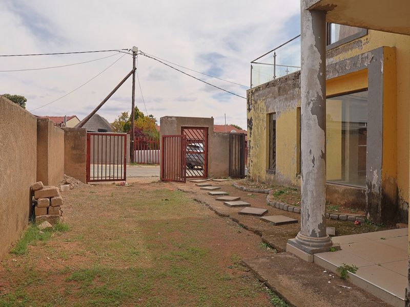 Front exterior view showing a gated entrance, paved walkway, and a building facade with visible wear and peeling paint on the structural column.
