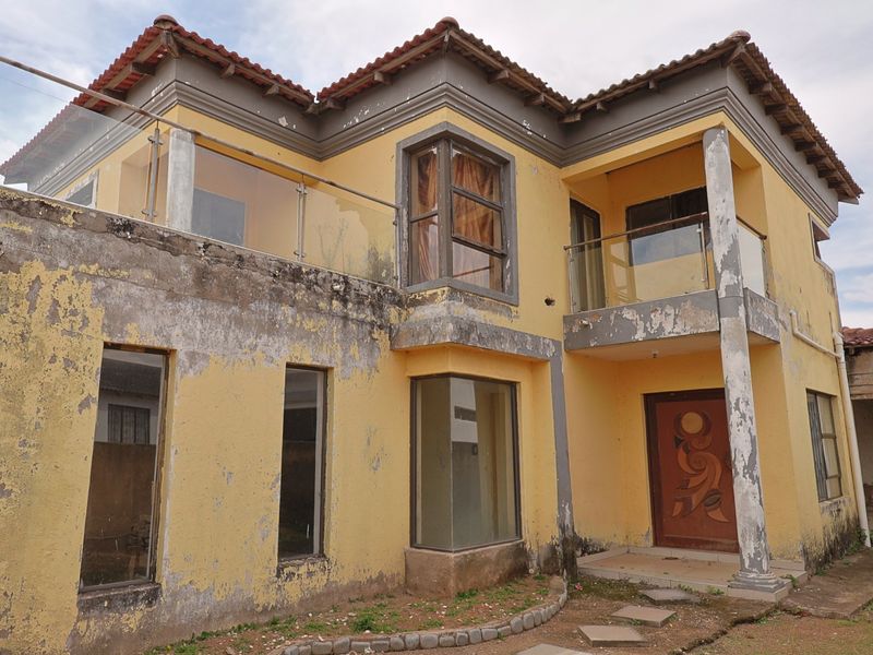 Two-story residential property featuring a yellow facade with glass balustrades and terracotta roof tiles, currently requiring significant exterior maintenance and repainting.
